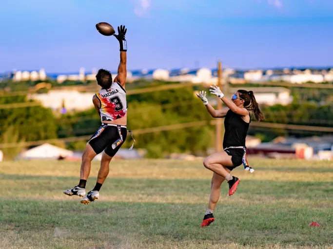 On a grassy practice field, an ATX Heat (Black) receiver in a white-and-black No. 3 jersey, gloves, and a flag belt leaps with one arm extended to snag a high pass while a defender in black athletic gear, gloves, sunglasses, and bright cleats jumps to contest. Cones mark the drill area with a cityscape in the distance under a clear sky.
