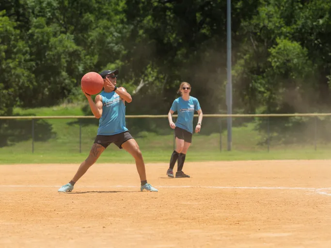 A kickball player in a blue jersey and visor is preparing to throw a red kickball on a dusty field, while another player in similar attire stands alert in the background.