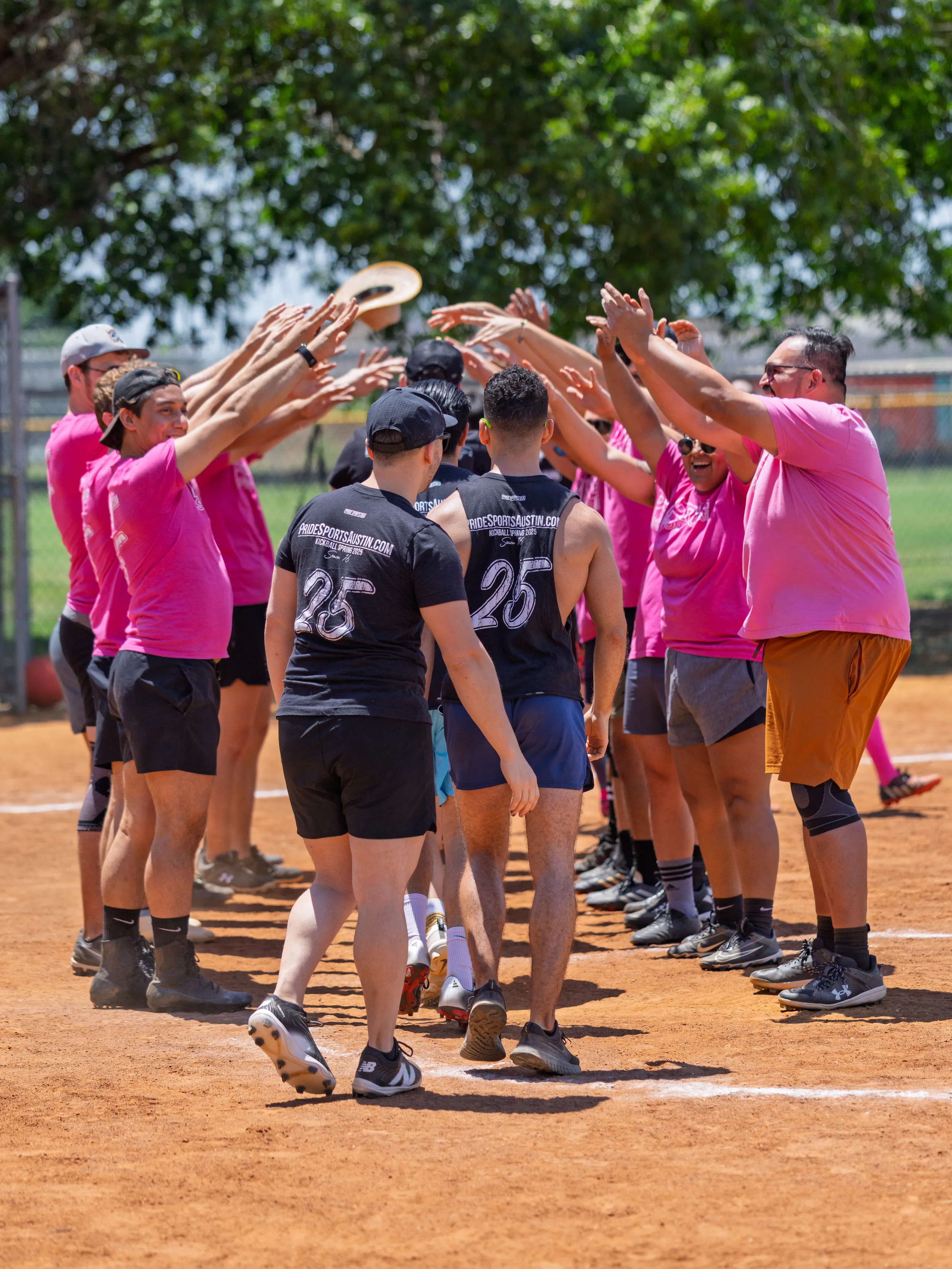 On a sunny Pride Kickball field, a team in bright pink shirts forms a raised-arm tunnel, cheering as players in black jerseys with numbers walk through. The dirt infield, athletic shoes, knee pads, and caps are visible, with green trees beyond the fence and one player holding a white cap overhead.