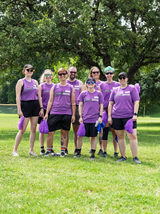 A group of eight kickball players wearing matching purple Stonewall Kickball shirts, shorts, and athletic shoes are standing together on a grassy field, holding purple pom-poms, with trees visible in the background.