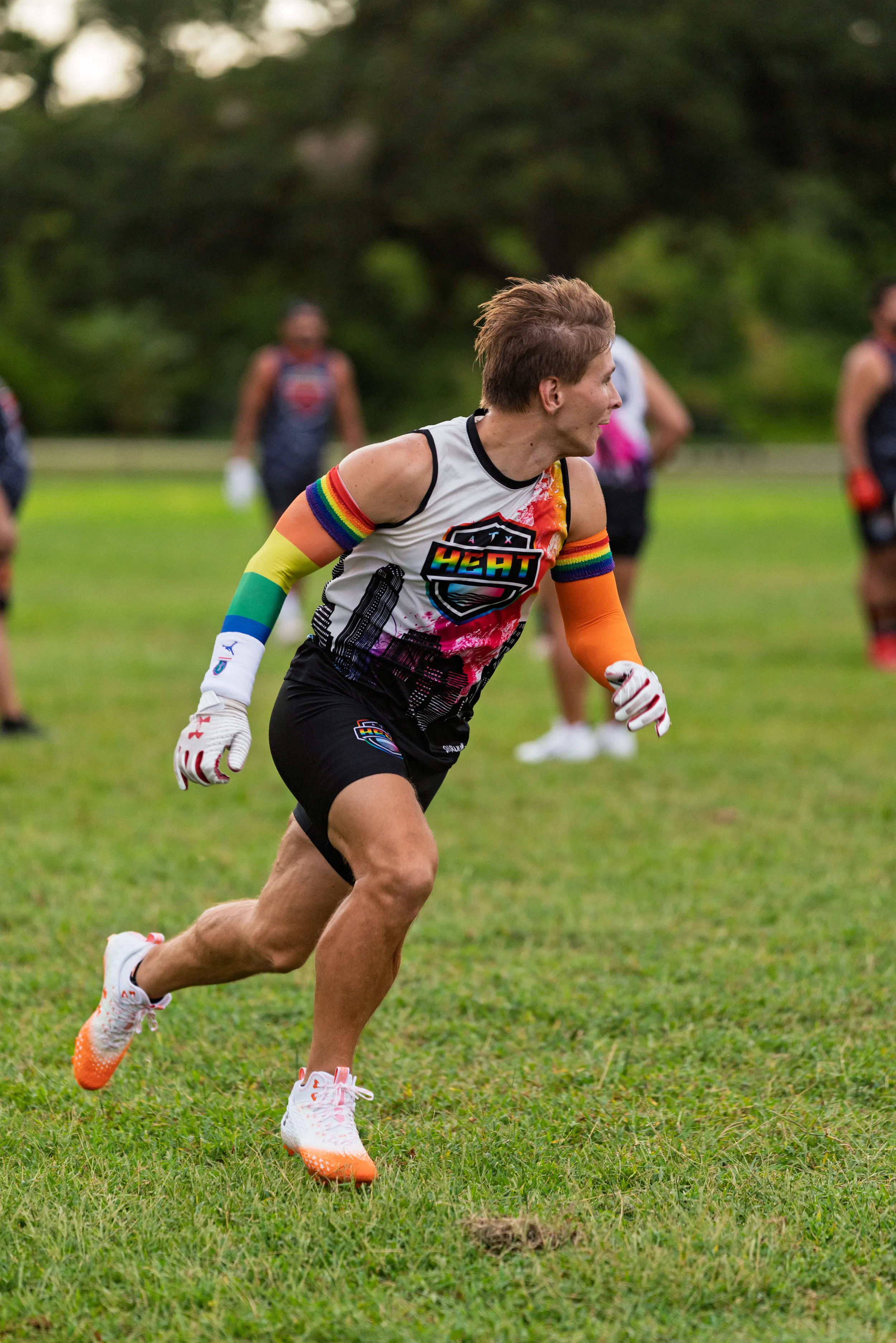 An ATX Heat (Orange) player cuts across a grassy field during flag football practice, looking to the side as if tracking a play. The athlete wears a sleeveless team jersey with bright graphics, black shorts, rainbow arm sleeves, white receiver gloves, and white cleats; blurred teammates and trees fill the background.