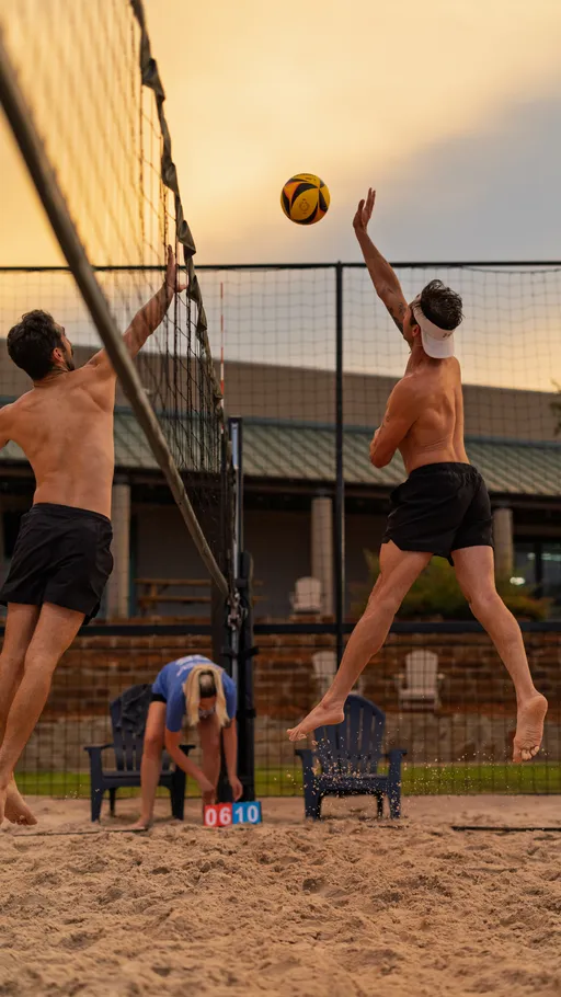 Two sand volleyball players in athletic shorts are mid-air at the net, one reaching up to block while the other jumps to spike the ball; a person in the background is adjusting the score on a scoreboard in a sandy outdoor court.