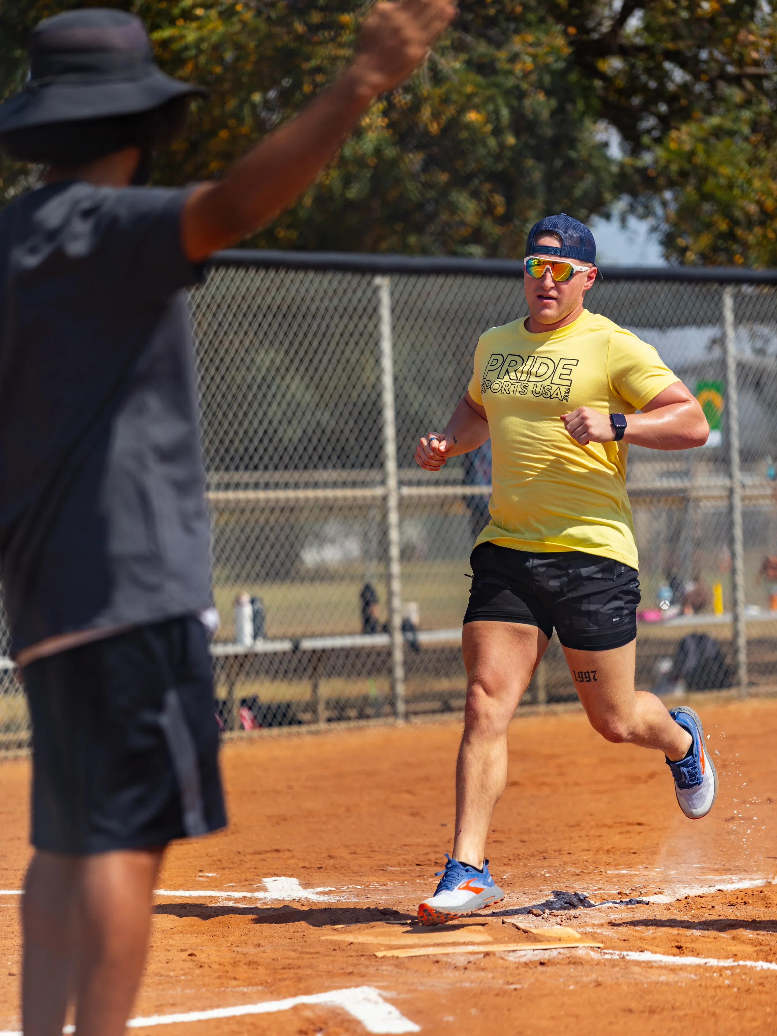 On a sunny kickball field, a player in a yellow “PRIDE SPORTS” shirt, black shorts, blue shoes, sunglasses, and a backward cap sprints across the home plate line, kicking up dust. A teammate or coach in a dark outfit and wide-brim hat raises both arms near the baseline, with a chain-link fence and trees in the background.