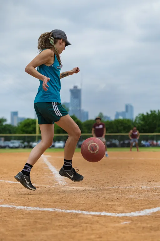 A player in athletic clothing and a cap is preparing to kick a red kickball on a dirt kickball field, with other players visible in the background near the bases.