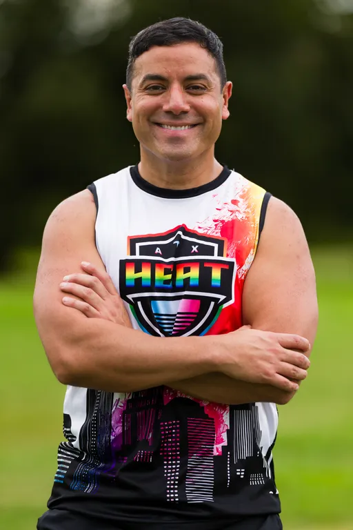 An ATX Heat player stands on a grassy practice field with arms crossed, wearing a sleeveless team jersey featuring a rainbow “HEAT” shield and abstract cityscape pattern, with blurred trees in the background during Gay Bowl 2025 preparations.
