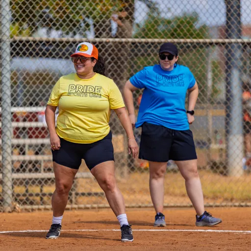 Two Pitch Please players stand on the kickball infield near a chain-link fence, pausing between plays. One wears a yellow “PRIDE” shirt, black shorts, and an orange cap; the other wears a blue “PRIDE” shirt, black shorts, and a black cap, both in athletic shoes.
