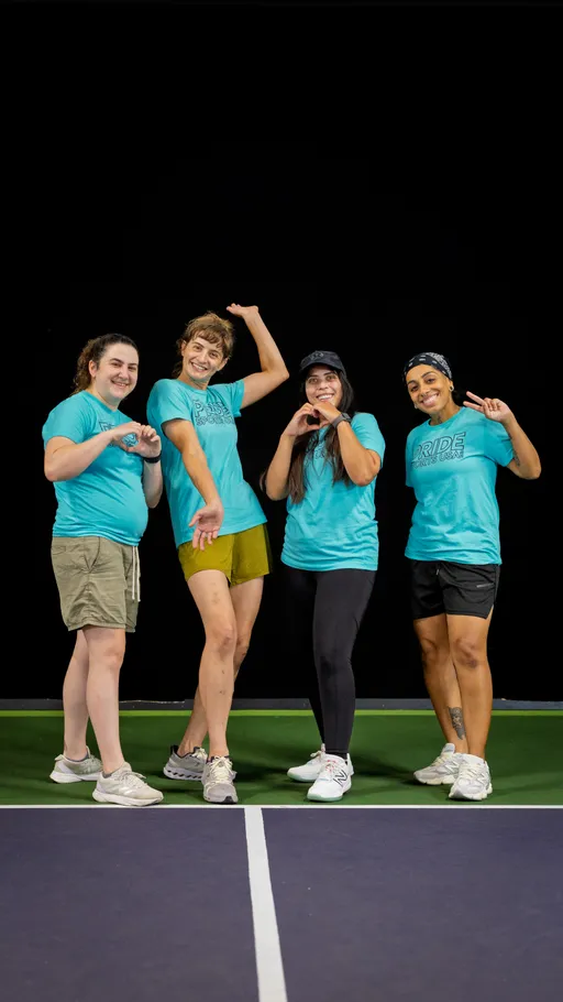 Four players wearing matching blue “Pride Pickleball” shirts pose cheerfully together on a brightly lit indoor pickleball court, making playful gestures and smiling.