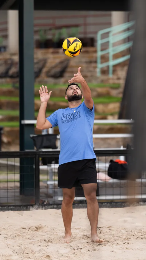 A player wearing a blue "Pride Sports" shirt and black shorts prepares to serve a yellow and black volleyball on a sand volleyball court.