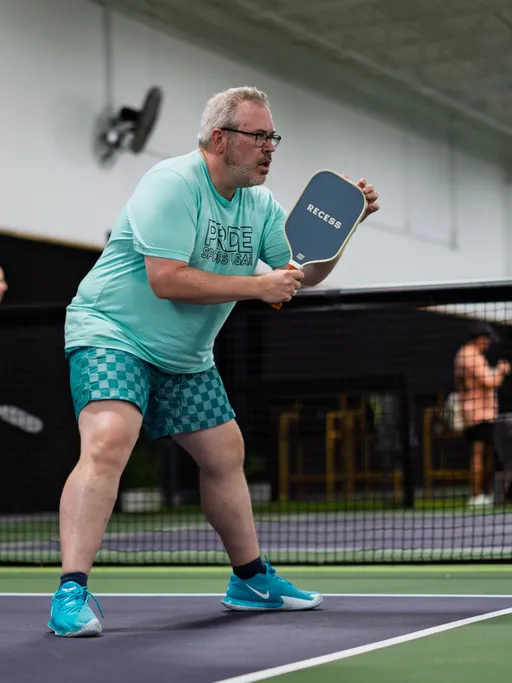 A pickleball player in a "Pride Sports League" shirt and checkered shorts prepares to return a shot with a paddle on an indoor court. The player is in an athletic stance, focused on the action, with a net and other participants visible in the background.