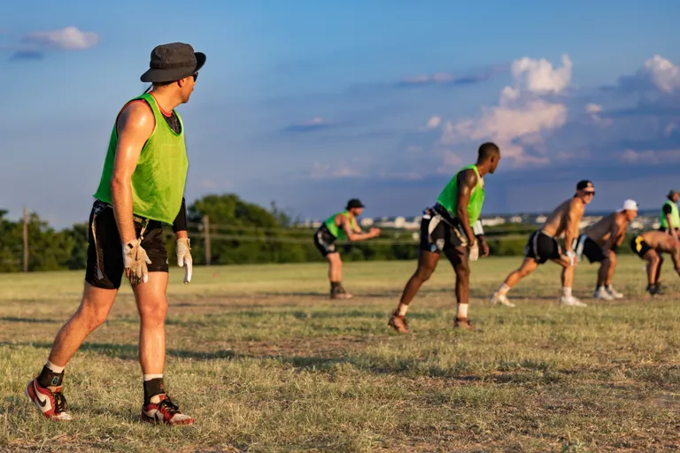 ATX Heat (Black) players practice flag football for Gay Bowl 2025 on a sunlit grassy field, spaced in a line and set in defensive stances. Athletes wear neon green pinnies, black shorts, flag belts, cleats, and gloves, with one player in a bucket hat, awaiting the snap during a drill.