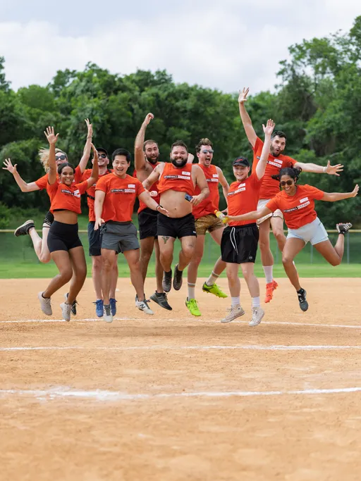 A group of kickball players wearing matching red shirts and athletic gear are jumping enthusiastically in the air on a sandy kickball field, with trees visible in the background.