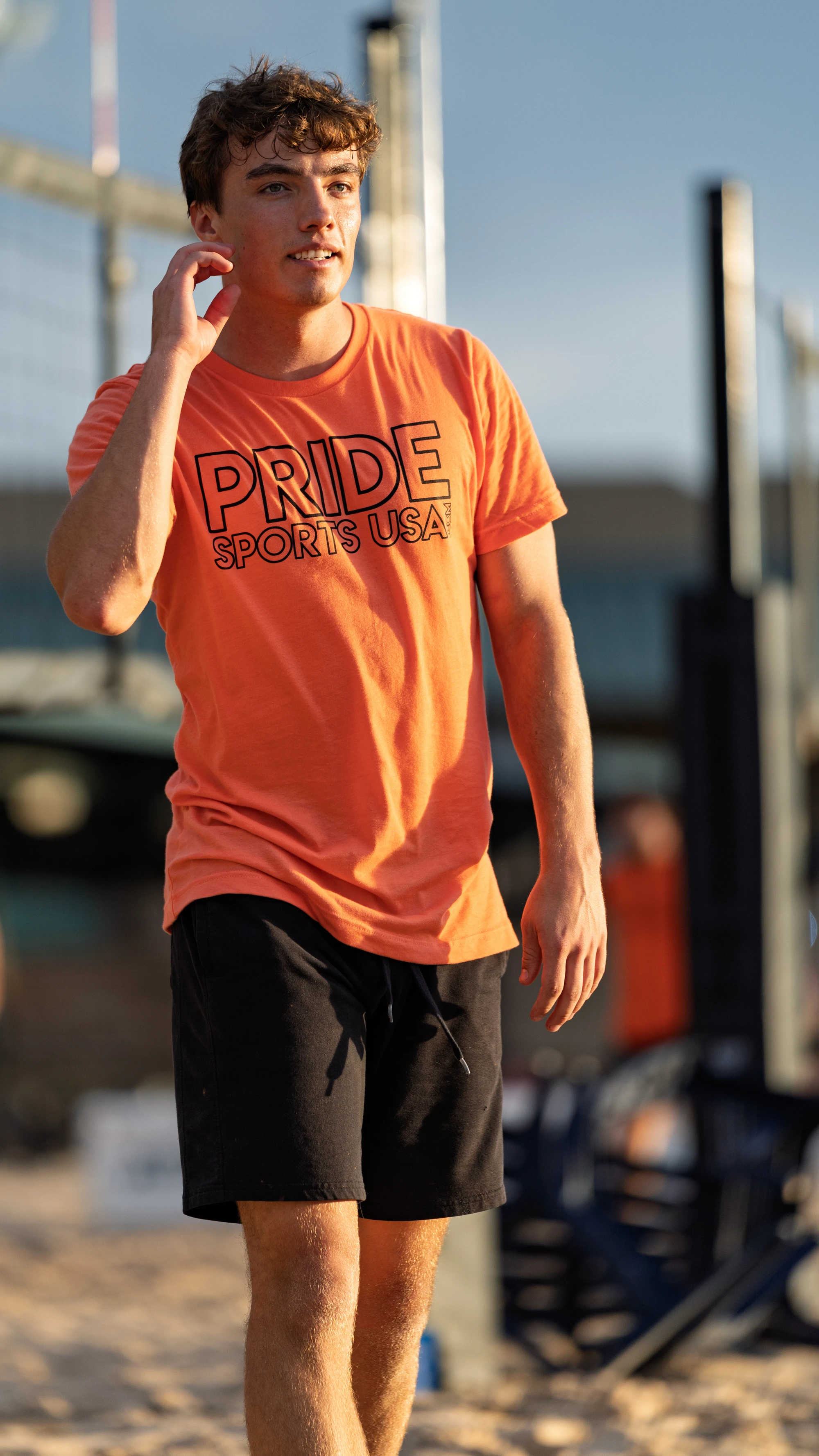 A volleyball player wearing a bright orange "Pride Sports USA" t-shirt and black shorts is walking on a sand court, with a volleyball net and equipment in the background.