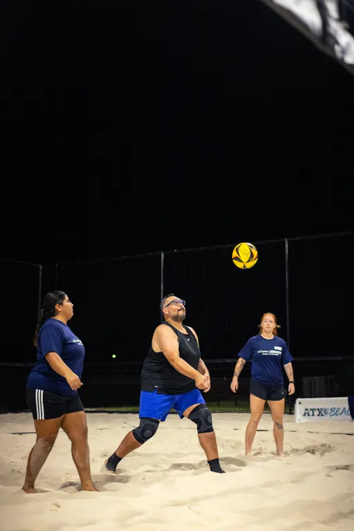Nighttime sand volleyball action: three players on a lit court watch a yellow-and-black ball descend. The central player, in a black tank and blue shorts with a knee brace and glasses, prepares a forearm pass while two teammates in navy shirts stand ready; fencing and an “ATX Beach” sign frame the background.