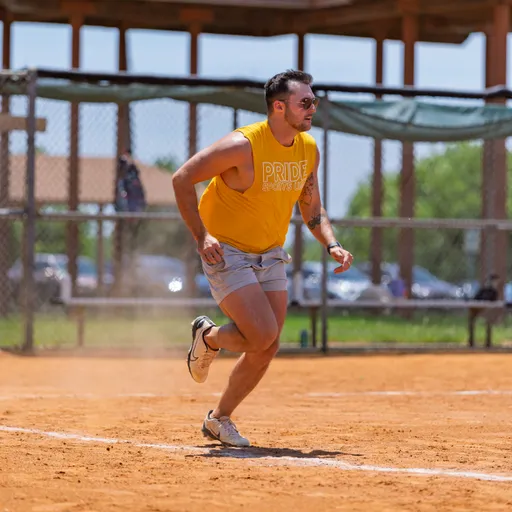 A player wearing a yellow "Pride Kickball" shirt, athletic shorts, and sneakers is running on a dusty kickball field, with a fence and shaded dugout area in the background.