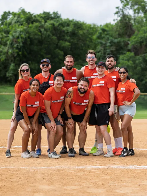 A group of kickball players wearing matching red team shirts pose together and smile on a dirt field, with trees visible in the background. Most players have their arms around each other, and some are crouched in front while others stand behind.