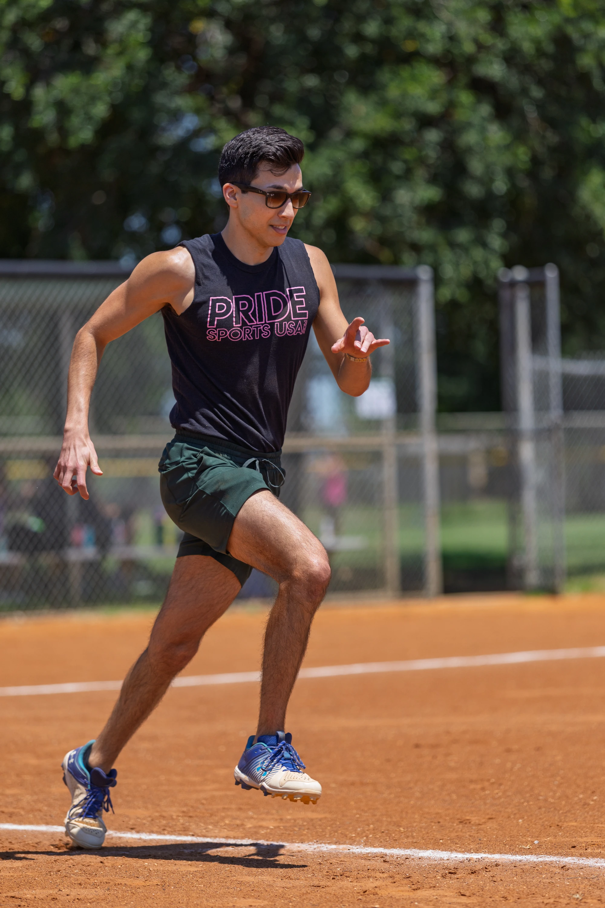 A player wearing sunglasses, a sleeveless "Pride Sports USA" shirt, shorts, and athletic shoes is running on a kickball field, with a chain-link fence and trees in the background.