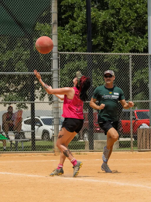 On a kickball field, a player in a pink shirt and black shorts is poised to catch a red kickball mid-air while another player in a green shirt and shorts runs nearby, both wearing athletic shoes and playing on a dirt field surrounded by a chain-link fence.