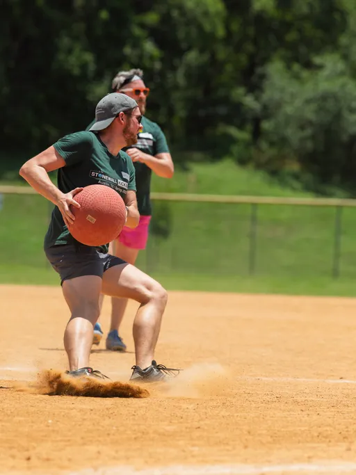 A kickball player wearing a green "Stonewall Kickball" shirt, shorts, and a backwards cap is about to throw a kickball on a sandy outdoor field, with another player in the background running and wearing bright shorts and sunglasses.