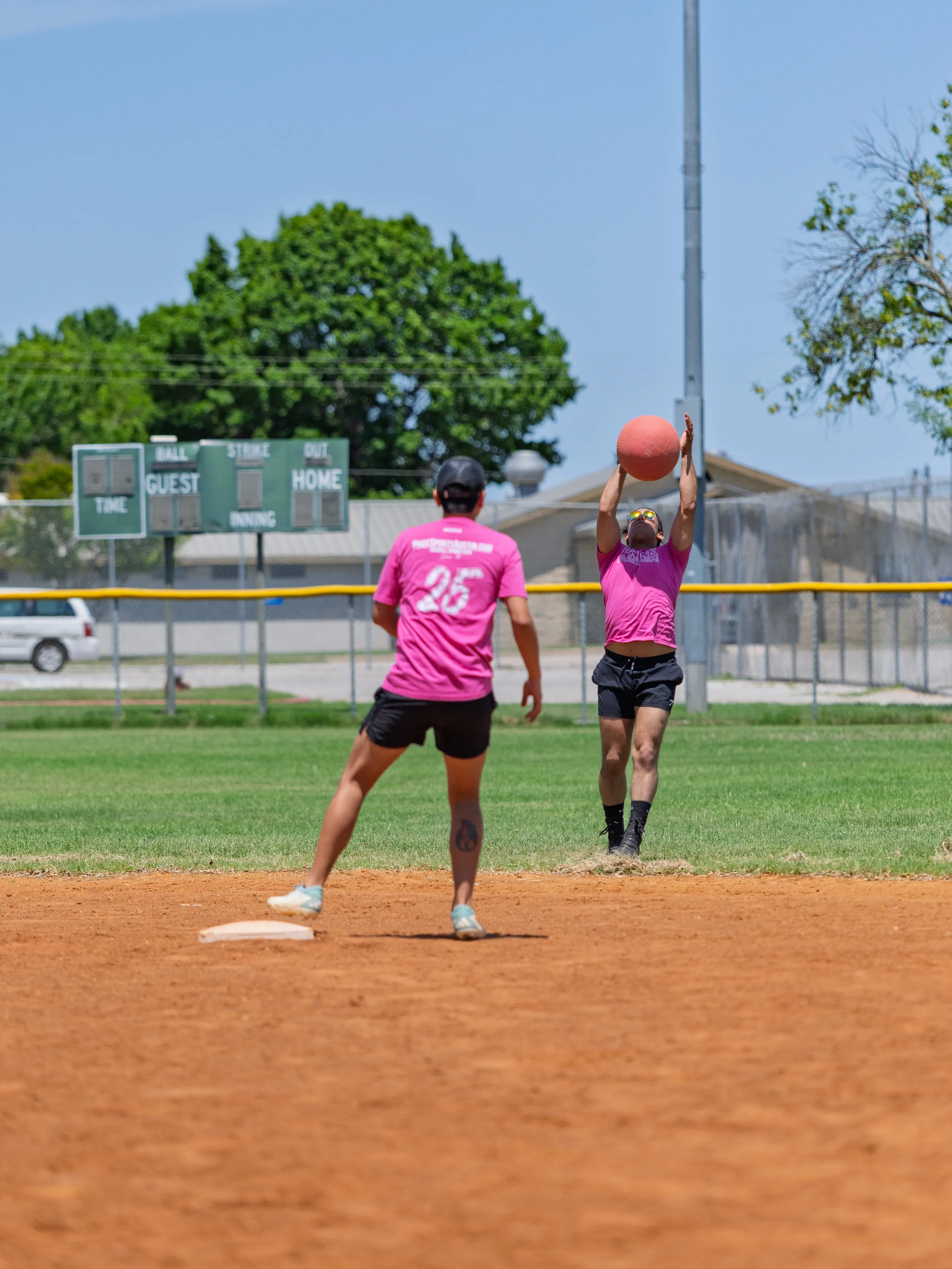 Two players in pink jerseys and black shorts are on a kickball field; one (jersey number 25) stands near a base while a teammate leaps with arms raised to catch a red kickball overhead near the pitcher’s area. A sunny outfield with a yellow fence and scoreboard sits in the background.