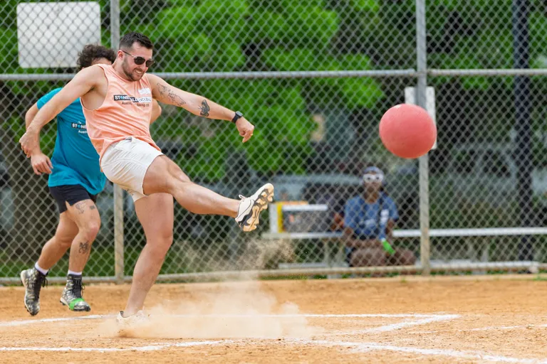 A player wearing sunglasses, a peach Stonewall Kickball tank top, and white shorts is kicking a red kickball on a dusty kickball field, with another player in a blue shirt running behind. There is a chain-link fence and some spectators in the background.