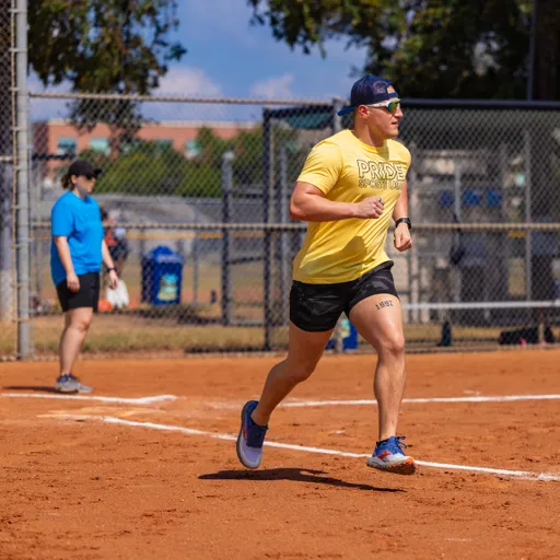 A player in a yellow Pride Kickball shirt, black shorts, blue sneakers, backward cap, and sunglasses sprints along the basepath on a sunny kickball field. A teammate in a blue shirt stands near the infield by the chain-link fence and trees.