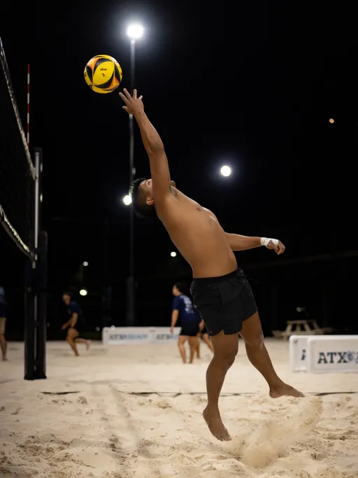 A player leaps beside the net on a lit sand volleyball court at night, arm fully extended to tip a yellow-and-blue ball as sand sprays from the jump. The athlete wears black shorts with taped fingers and wrist; teammates and opponents in dark jerseys stand in the background near barriers labeled “ATX.”
