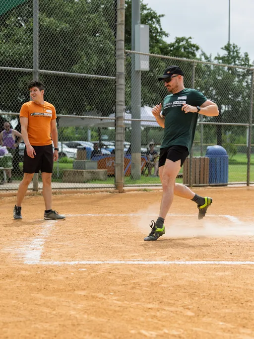 One player in a green shirt, black shorts, and a cap is running across home plate, kicking up dust, while another player in an orange shirt watches from nearby on a kickball field enclosed by a chain-link fence.