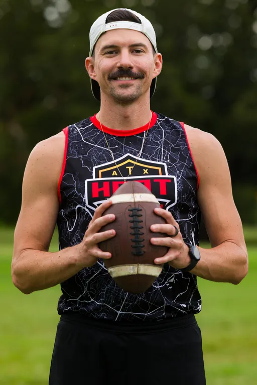 An ATX Heat player stands on a grassy field facing forward, gripping a football with both hands at chest level during practice for Gay Bowl 2025. The player wears a black sleeveless ATX Heat jersey with red trim, a light cap worn backward, a wristwatch, and a thin necklace, with trees softly out of focus behind.