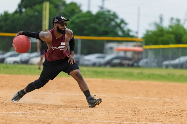 A kickball player wearing a maroon jersey, black athletic gear, and a cap is in mid-action, preparing to throw a red kickball on a sandy field with a fence and parked cars in the background.