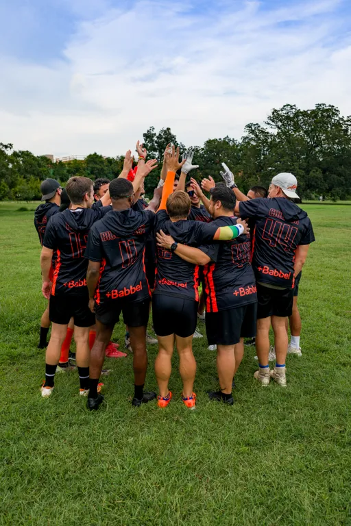 On a grassy park field, ATX Heat (Orange) players huddle with arms raised in a team cheer during flag football practice for Gay Bowl 2025. They wear matching black jerseys with orange accents and numbers, with several players in gloves, caps, and arm sleeves; trees and a cloudy sky fill the background.