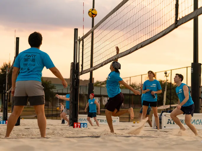 During a Stonewall Sand Volleyball match at sunset, a player in a blue team shirt leaps at the net to tip a yellow‑and‑black ball while teammates in matching blue shirts and shorts ready for the play; sand sprays up, with the scoreboard (17–09), fencing, and courts in the background.