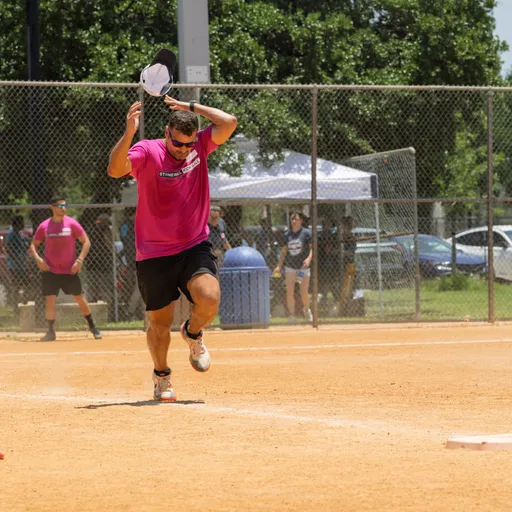 A player wearing a bright pink shirt and black shorts is mid-stride on a kickball field, holding a cap above their head, with other players and spectators visible near the fence in the background.