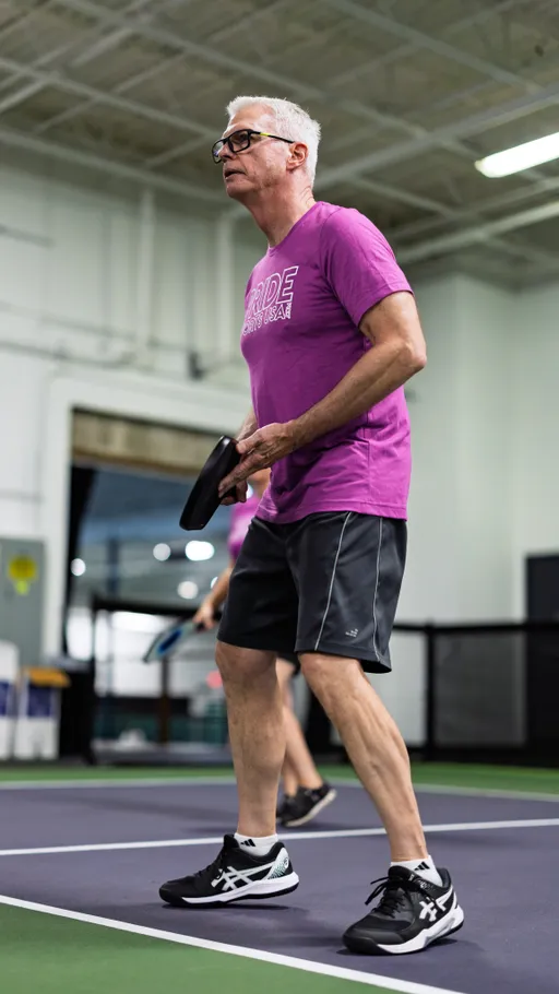 A pickleball player wearing a "Pride Pickleball" t-shirt, athletic shorts, and sneakers is preparing to serve on an indoor pickleball court, holding a paddle and focusing on the game. Another player is visible in the background, also engaged in play.