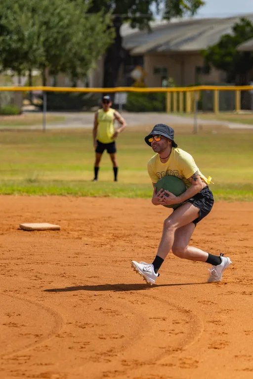 On a sunny kickball field, a player in a yellow jersey, black shorts, bucket hat, and sunglasses charges across the infield while cradling a green kickball near a base. Another player in matching colors stands in the outfield by a yellow-topped fence, watching the play.