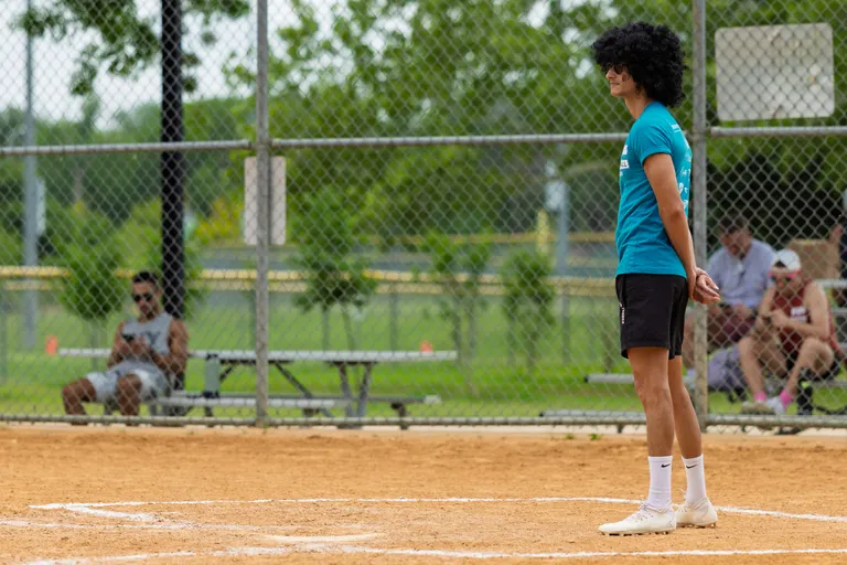A kickball player in a teal shirt, black shorts, and white athletic shoes stands on the field near home plate, facing forward with hands behind their back while spectators watch from benches behind a chain-link fence.