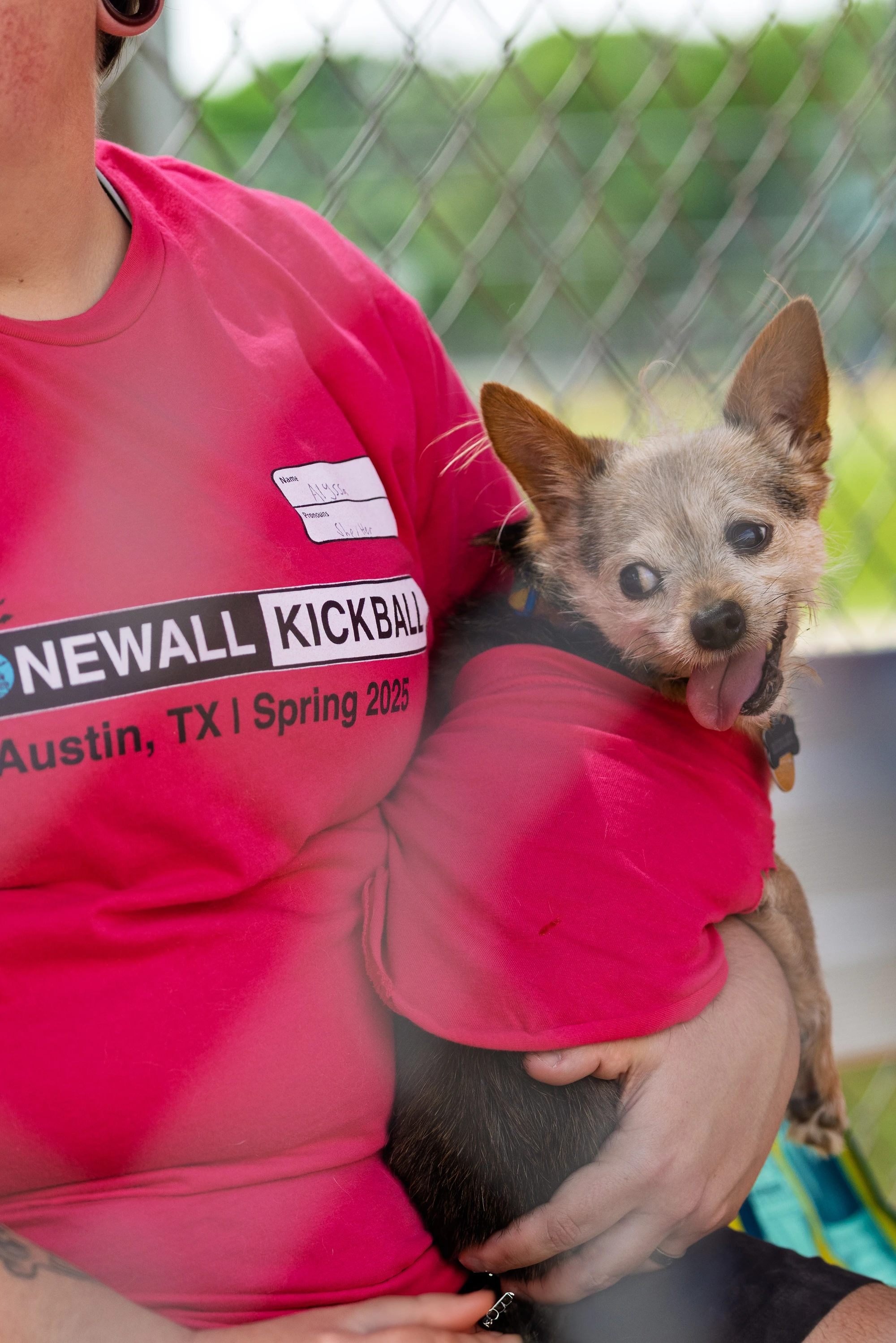 A player wearing a bright red "Stonewall Kickball - Austin, TX - Spring 2025" shirt is sitting and holding a small dog wrapped in a matching red blanket, with a chain-link fence and greenery in the background.
