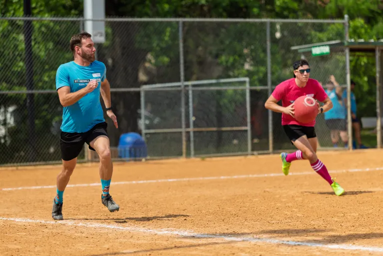 Two players are in action on a kickball field; one player in a blue shirt and matching socks is running while another player in a pink shirt, striped knee socks, and sunglasses prepares to throw a large red kickball. They are surrounded by a fenced outdoor field with trees in the background.