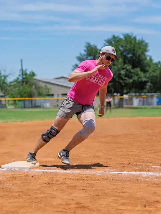 A kickball player wearing a pink "Pride Sports League" shirt, gray shorts, hat, sunglasses, and knee pads is running around a base on a sunny outdoor field.