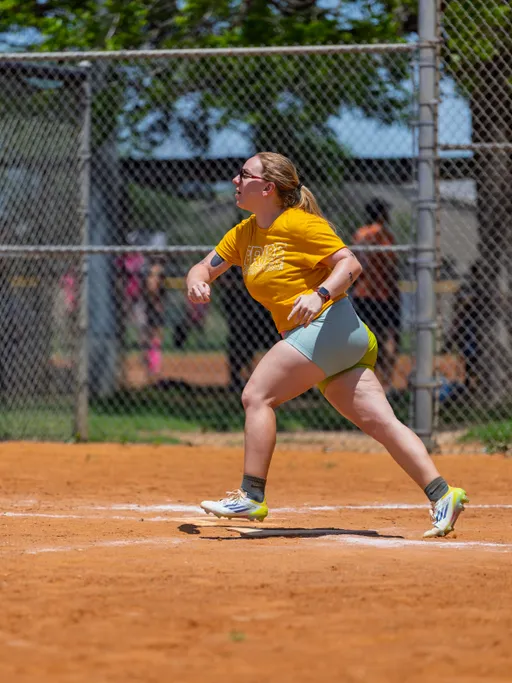A player wearing a yellow shirt, light shorts, and athletic shoes is mid-stride on a sandy kickball field, appearing to have just kicked the ball during a game, with a chain-link fence and other people visible in the background.