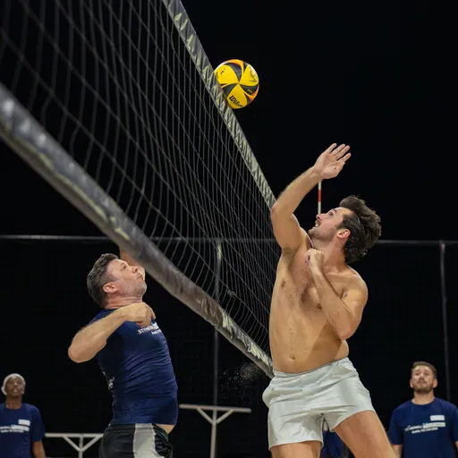On a lit sand volleyball court at night, a player in white shorts jumps at the net to tip or block a yellow-and-black volleyball near the antenna while an opposing player in a navy shirt reaches up from the other side. Teammates in matching shirts watch from the background as the rally unfolds.