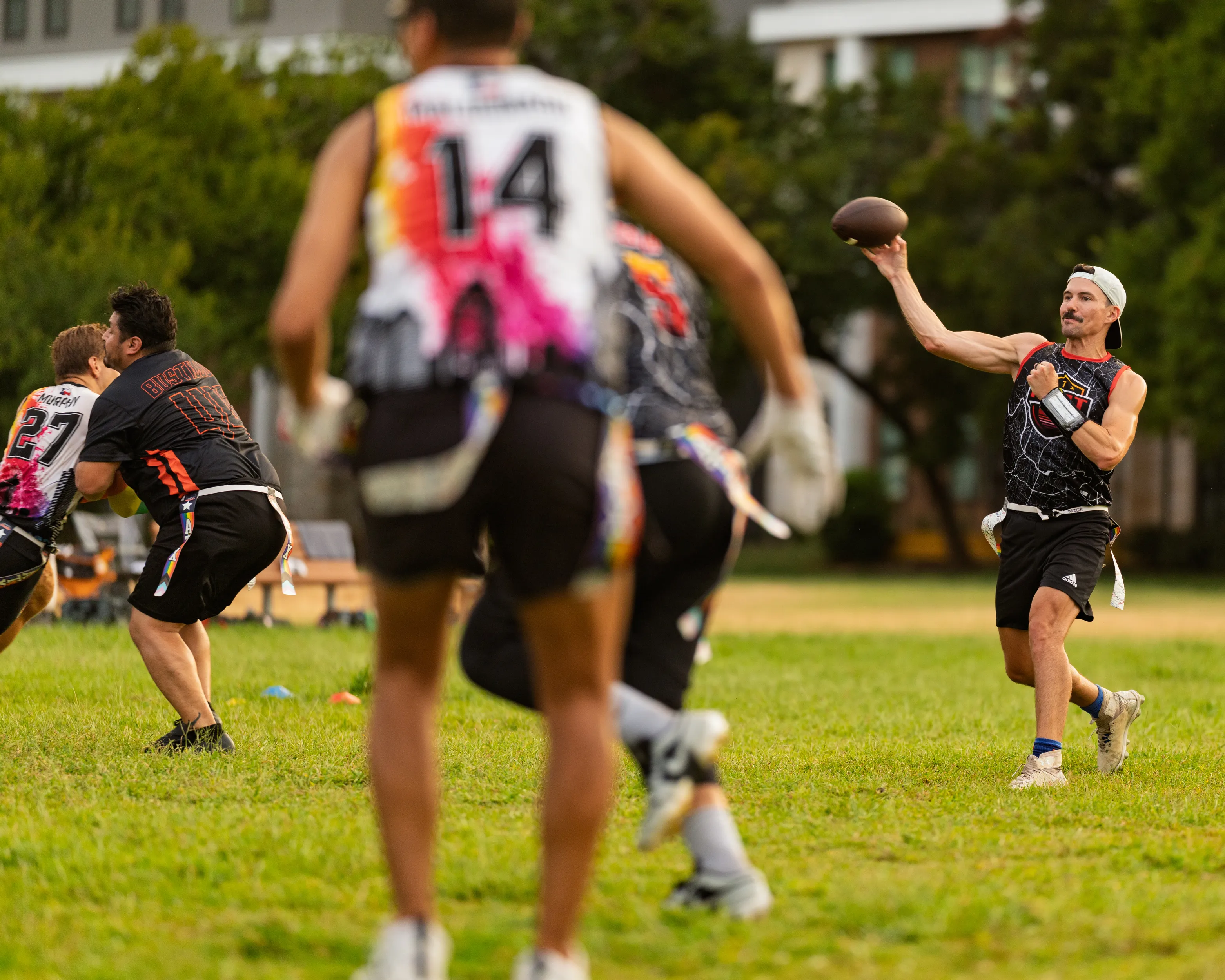 On a grassy park field, ATX Heat (Orange) practices for Gay Bowl 2025: a quarterback in a white cap throws a football while teammates in black jerseys with bright orange and pink patterns and numbered backs run routes, their flag belts visible. Cones and benches line the sideline with trees and buildings in the background.