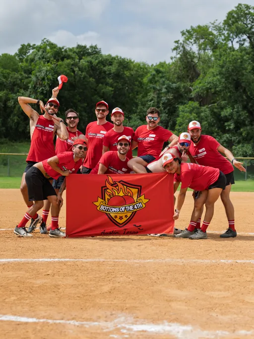 A group of kickball players poses together on a kickball field, all wearing matching red shirts, hats, and socks, smiling and gathering around a team banner that reads "Bottoms of the 4th, Austin, Texas."