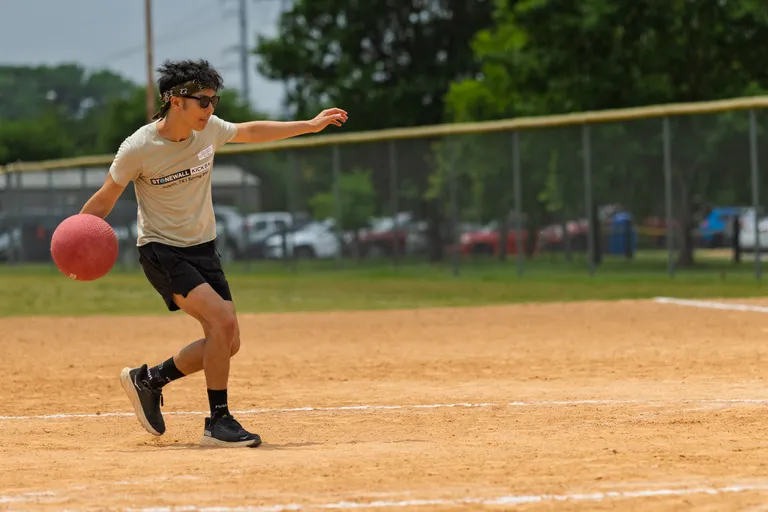 A kickball player wearing a light t-shirt, black shorts, black athletic shoes, sunglasses, and a headband is preparing to kick a red kickball on a dirt field, with a fence and trees in the background.