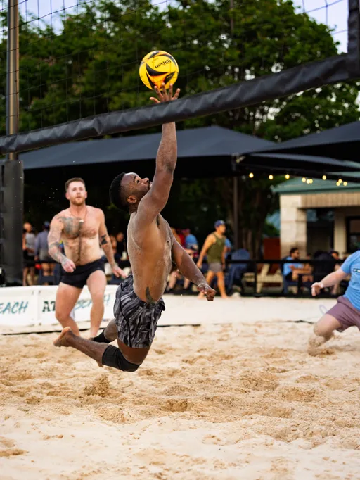 A volleyball player in athletic shorts jumps mid-air under a sand volleyball net, reaching to hit the ball with one hand, while other players watch and move nearby on a sand court with trees and spectators in the background.