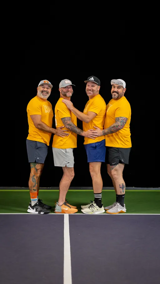 Four pickleball players wearing matching yellow shirts, shorts, hats, and sneakers are standing in a row on a court, each with their arms around the person next to them and smiling toward the camera. The background is a dark, solid color, highlighting the group’s team spirit and camaraderie.