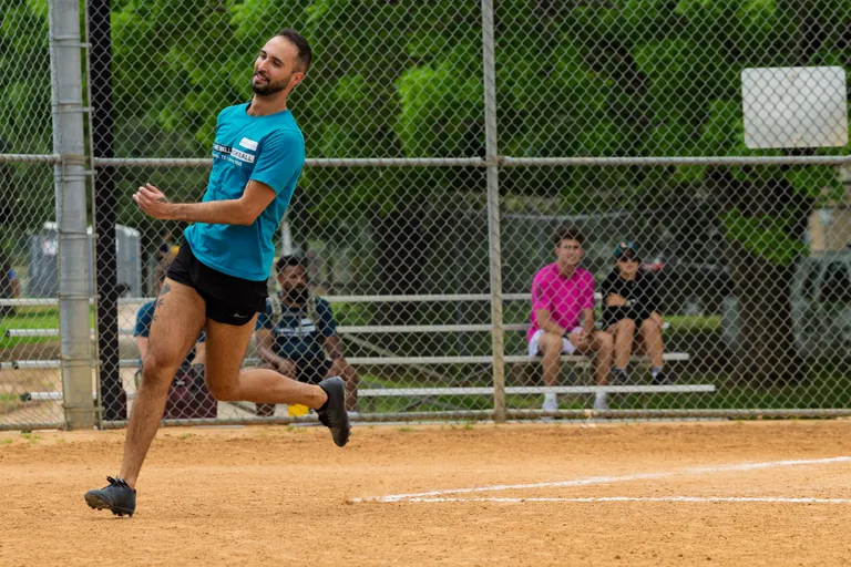 A player wearing a teal team shirt, black shorts, and cleats is running on a kickball field, mid-action, with spectators seated on bleachers behind a chain-link fence in the background.