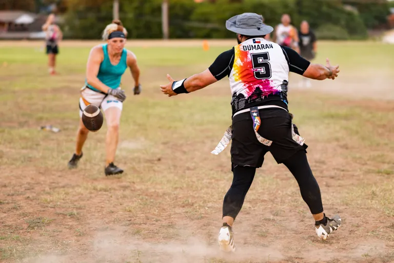 During ATX Heat (Black) practice for Gay Bowl 2025, a ball carrier in a teal top and headband runs across a dusty flag football field while a defender in a black multicolor jersey and wide-brim hat charges with arms out to grab the hip flags; blurred teammates and orange cones dot the background.