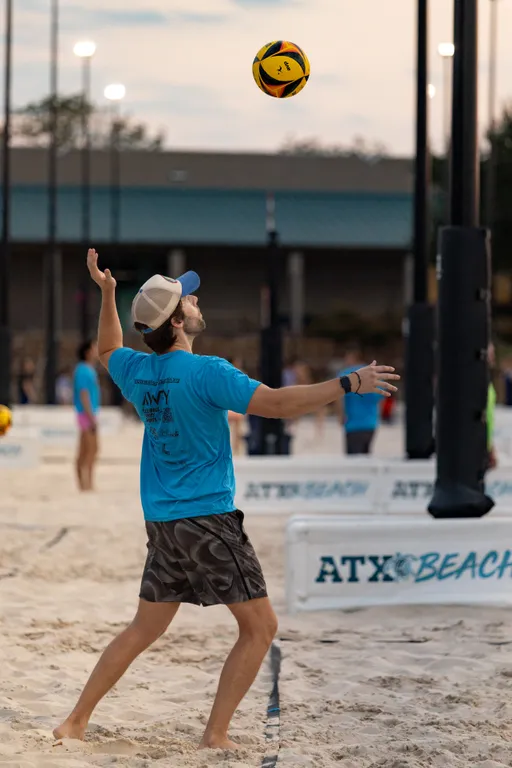 A player in a bright blue team shirt, patterned shorts, and a trucker-style cap tosses a yellow-and-black volleyball overhead, preparing to serve on a sand volleyball court at dusk while wearing a wristband and holding a small red cup. Multiple courts and players are active in the background under field lights, with “ATX Beach” signage on the court barriers.