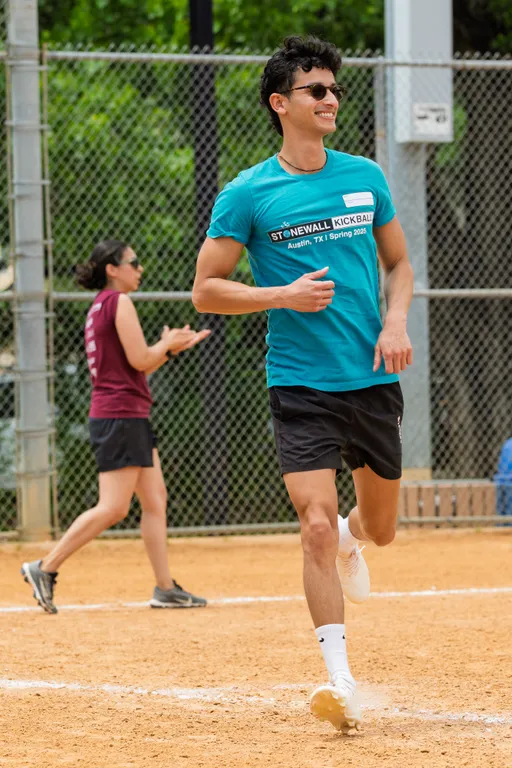 A player in a blue "Stonewall Kickball" shirt and black shorts is running on a kickball field, smiling, while another player in a maroon shirt and black shorts is visible in the background. The scene takes place on a sandy field with a chain-link fence and greenery behind it.