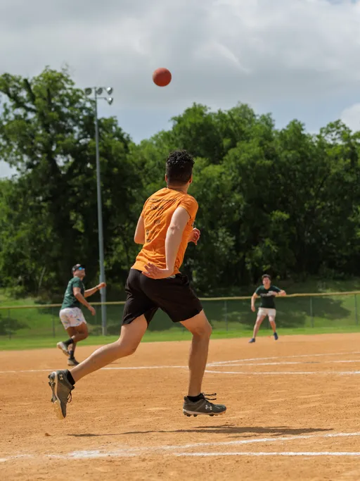 A player in an orange shirt and black shorts runs toward a kickball in mid-air on a dirt field, while other players in green shirts prepare to catch or field the ball, with trees and a fence visible in the background.